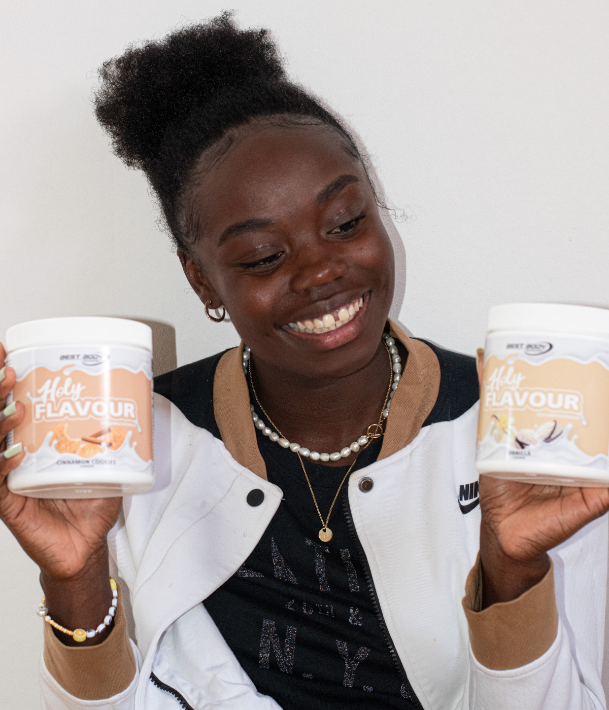 Junge Frau mit dunklem Lockenzopf lächelt und hält zwei Dosen Best Body Nutrition Holy Flavour Pulver in den Händen.