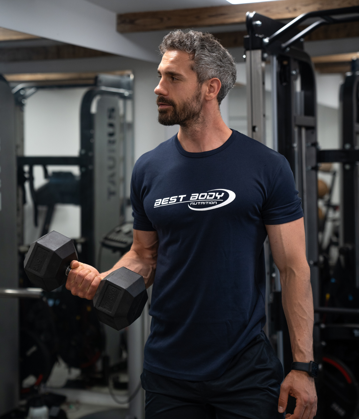 Mann mit grauen Haaren trainiert im Fitnessstudio und hält eine schwarze Kurzhantel, er trägt ein dunkelblaues T-Shirt mit Best Body Nutrition Logo.