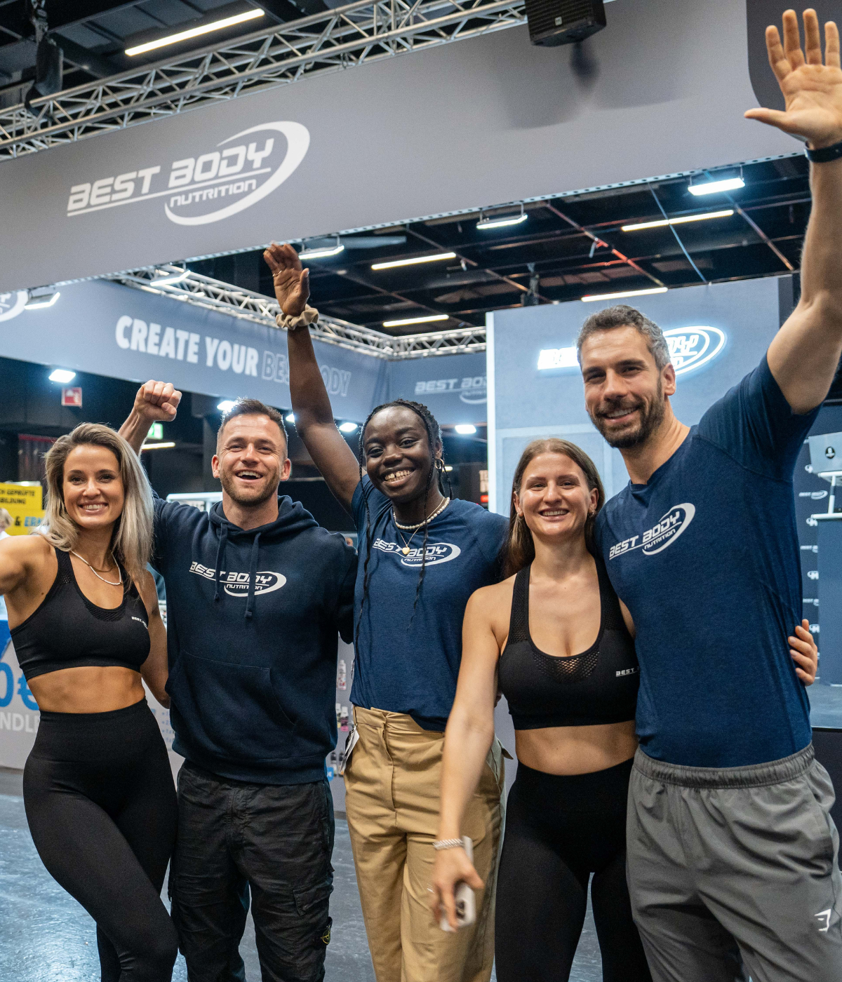 Fünf gut gelaunte Menschen stehen vor dem Messestand von Best Body Nutrition und posieren lachend mit erhobenen Armen.