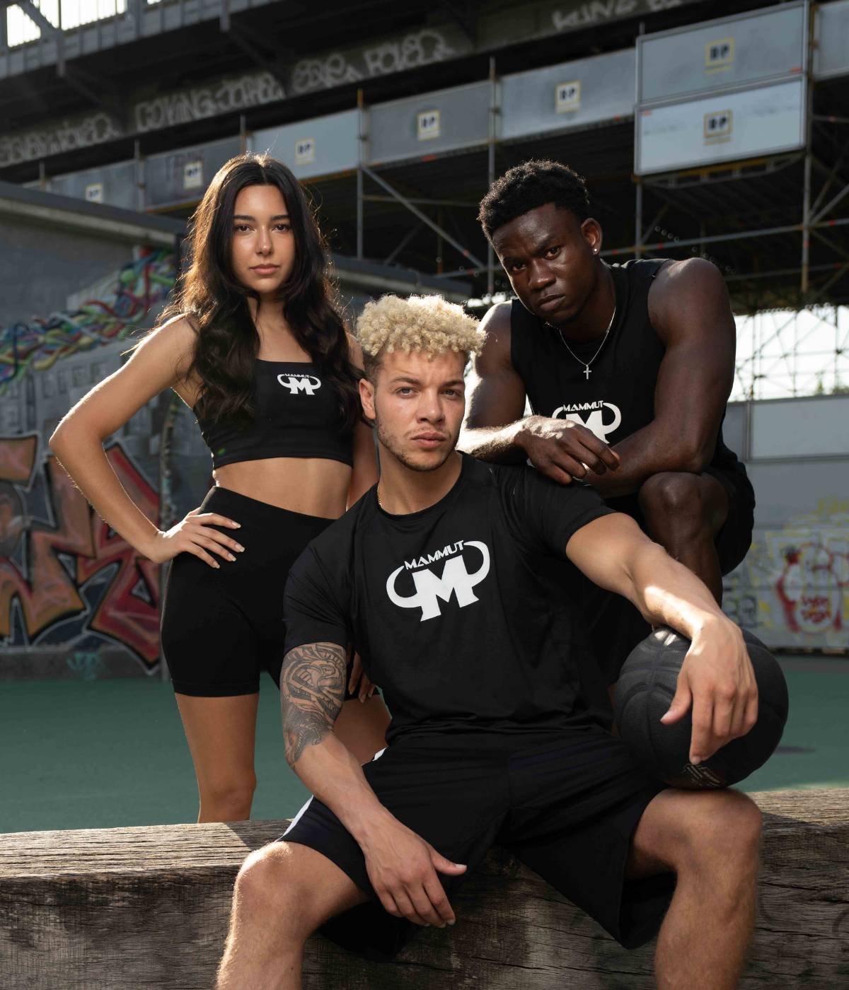 Zwei junge Männer und eine junge Frau in schwarzer Mammut Nutrition Sportkleidung posieren auf einem Basketballplatz mit Graffiti-Wänden.
