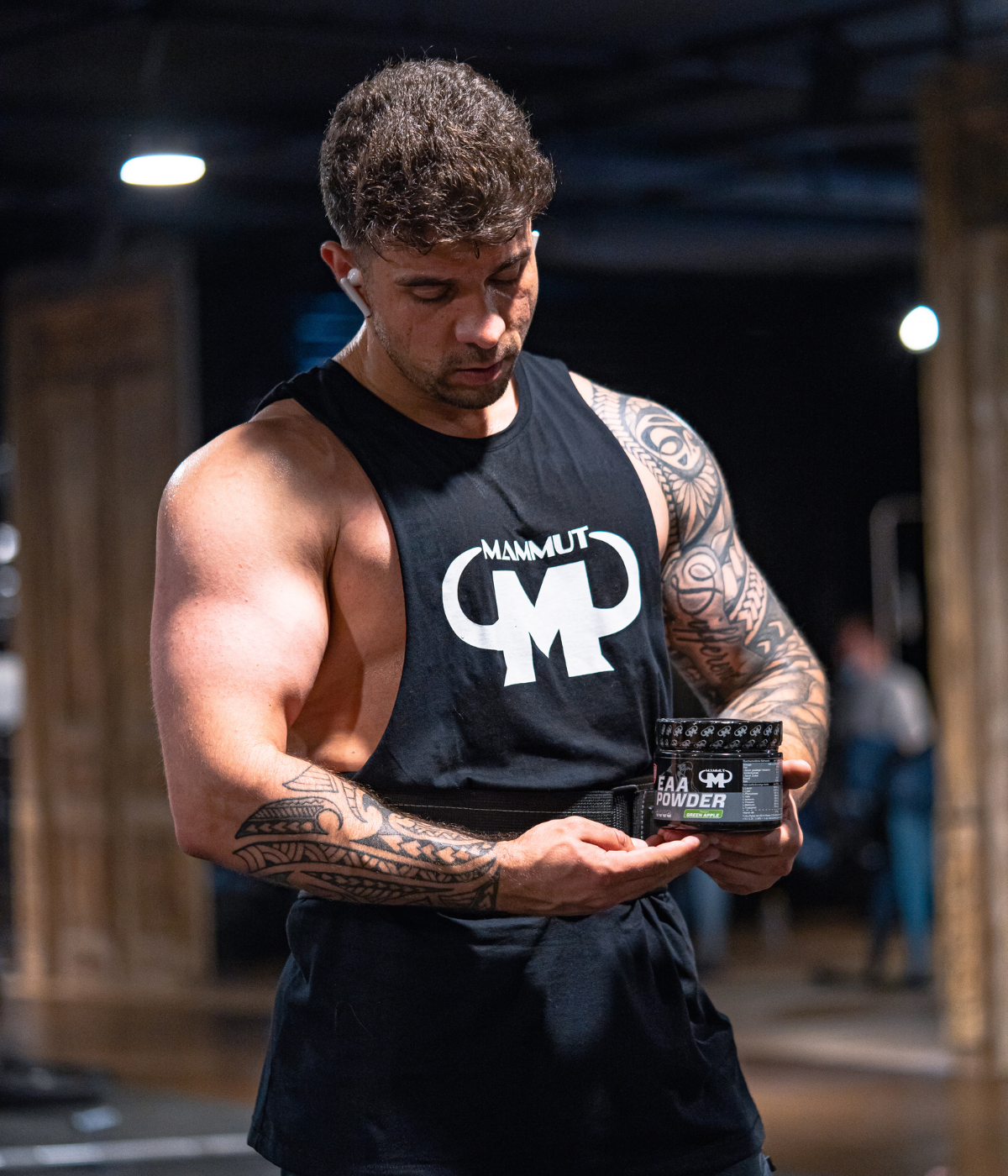 Muskulöser Mann im schwarzen Mammut Nutrition Tanktop steht im Fitnessstudio und hält eine Dose Mammut Nutrition EAA Powder in den Händen.