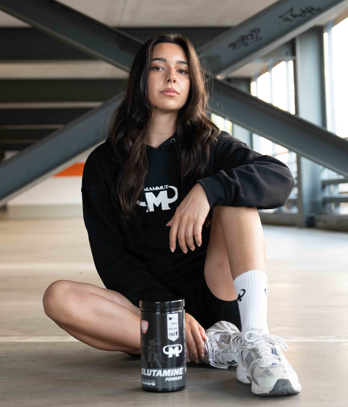 Junge Frau im schwarzen Mammut Nutrition Hoodie sitzt in einer Halle und hat eine Dose Mammut Nutrition Glutamine vor sich auf dem Boden stehen.