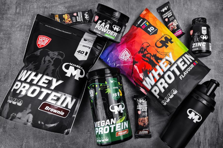 Sortiment von Mammut Nutrition Produkten, darunter Whey Protein, Vegan Protein, EAA Powder, Crunchy Protein Bars, Zink-Kapseln und ein schwarzer Shaker, auf grauem Hintergrund arrangiert.