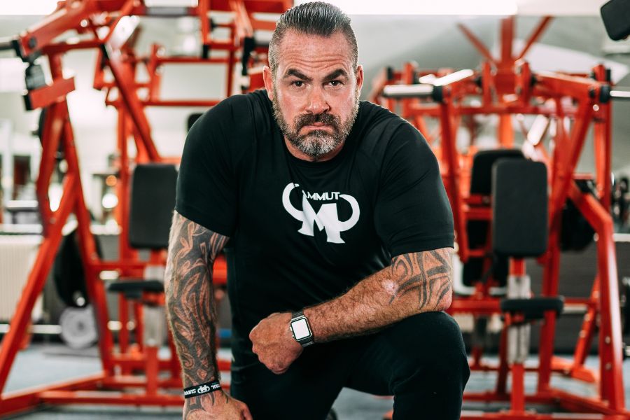 Mann mit tätowierten Armen trägt ein schwarzes Mammut Nutrition T-Shirt und kniet ernst blickend in einem Fitnessstudio mit roten Trainingsgeräten.