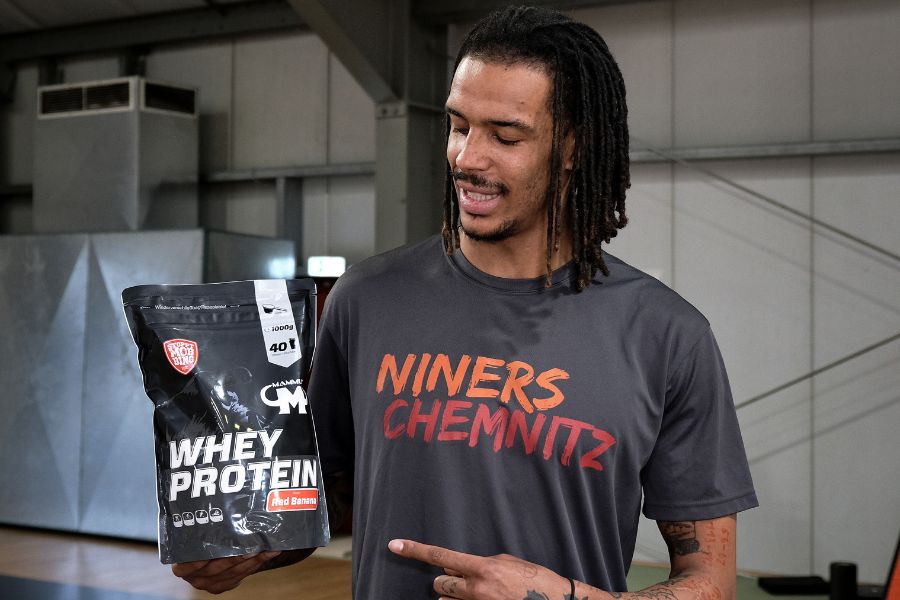 Basketballspieler im Niners Chemnitz Shirt hält eine Packung Mammut Nutrition Whey Protein in der Sorte Red Banana und zeigt mit dem Finger darauf.