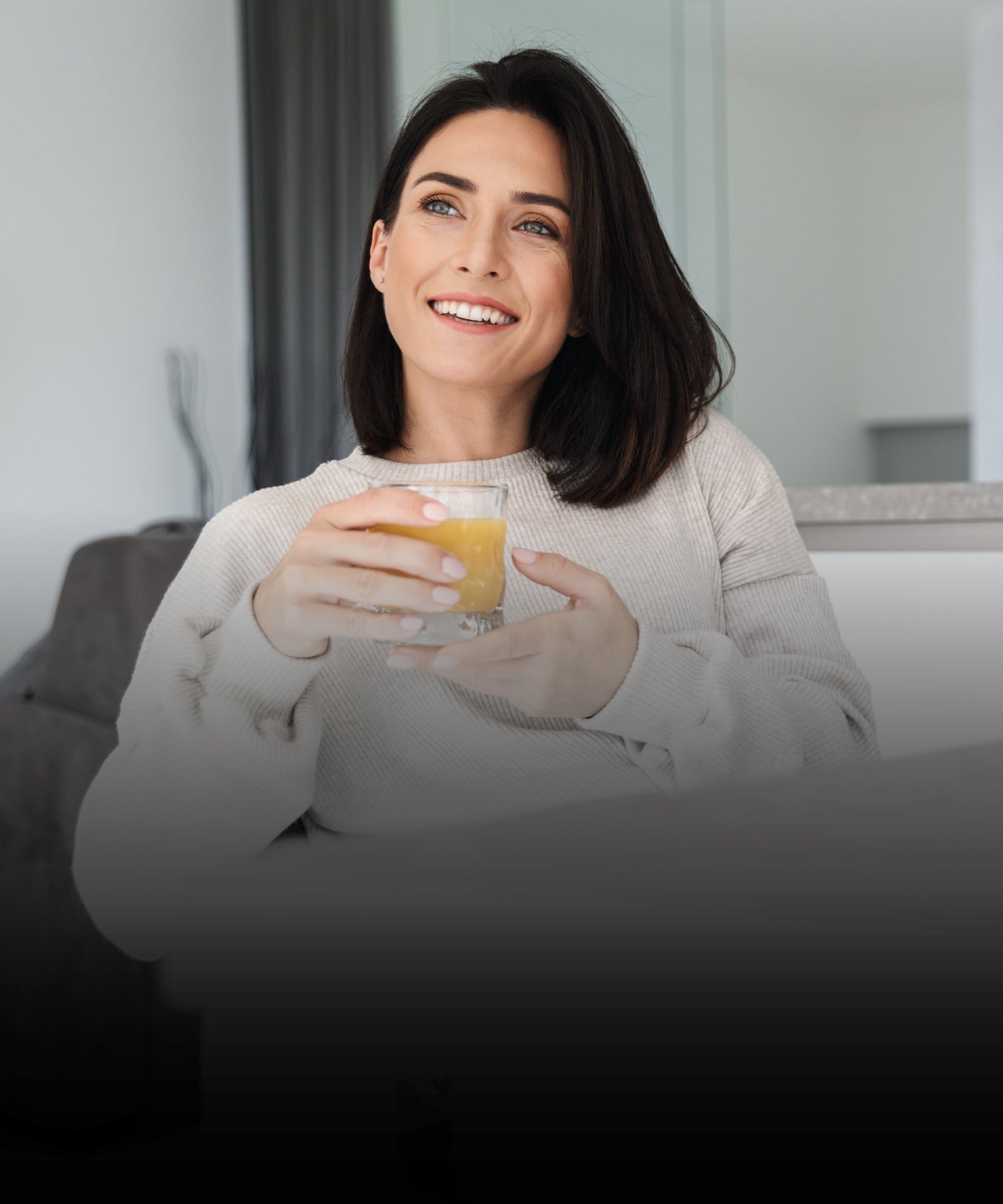 Frau mit schulterlangem dunklem Haar sitzt entspannt in heller Wohnung und lächelt, während sie ein Glas mit orangem Getränk in den Händen hält.