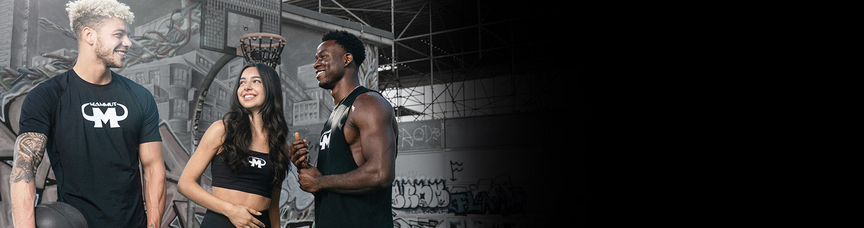 Drei sportlich gekleidete junge Menschen stehen lachend in einer Halle mit Basketballkorb und Graffiti-Wänden.